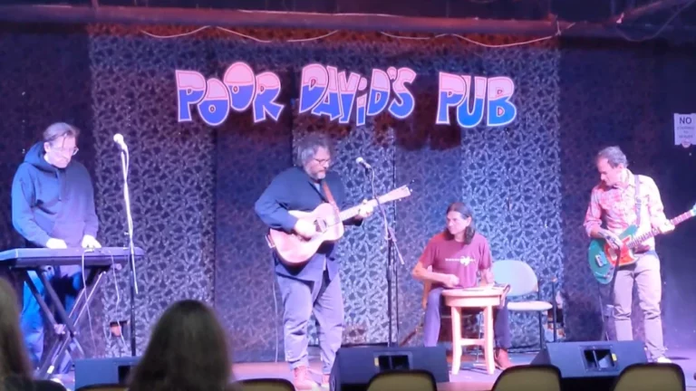 Musicians performing during open mic night at Poor David's Pub, featuring a guitarist, keyboard player, and percussionist on stage