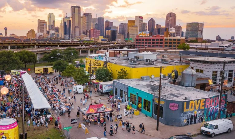 Aerial view of a crowded outdoor event during Memorial Day weekend in Dallas, with food trucks, vendor tents, and the city skyline in the background at sunset