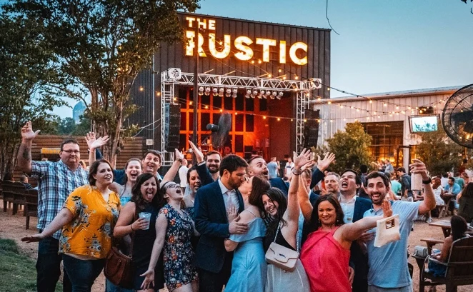 Group of people enjoying a lively evening at The Rustic, a bar in Dallas, with a stage and outdoor seating in the background.