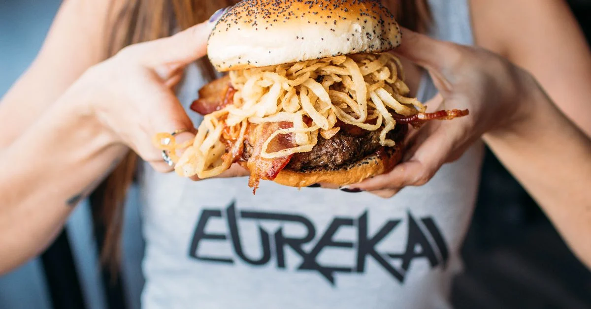 Eureka Cowboy Burger with crispy onions, bacon, and a beef patty held by a person wearing a Eureka t-shirt