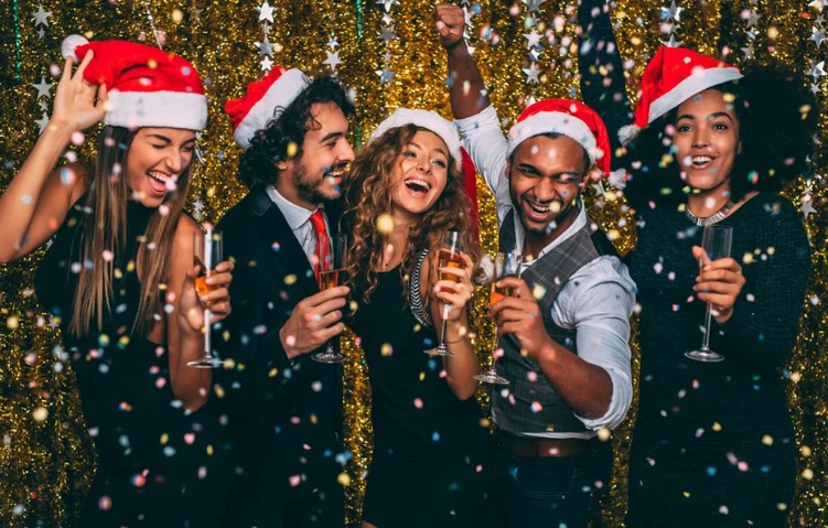 Group of friends wearing Santa hats celebrating at a festive party with confetti and drinks against a gold sequin backdrop