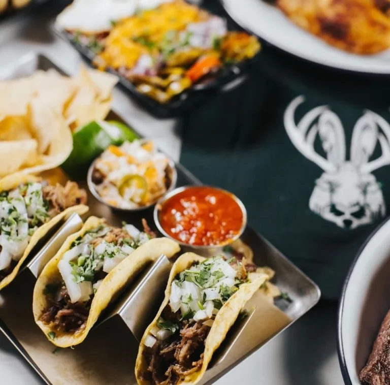 A tray of three tacos topped with cilantro and onions, served with a side of salsa, chips, and a jalapeño, with other dishes in the background.
