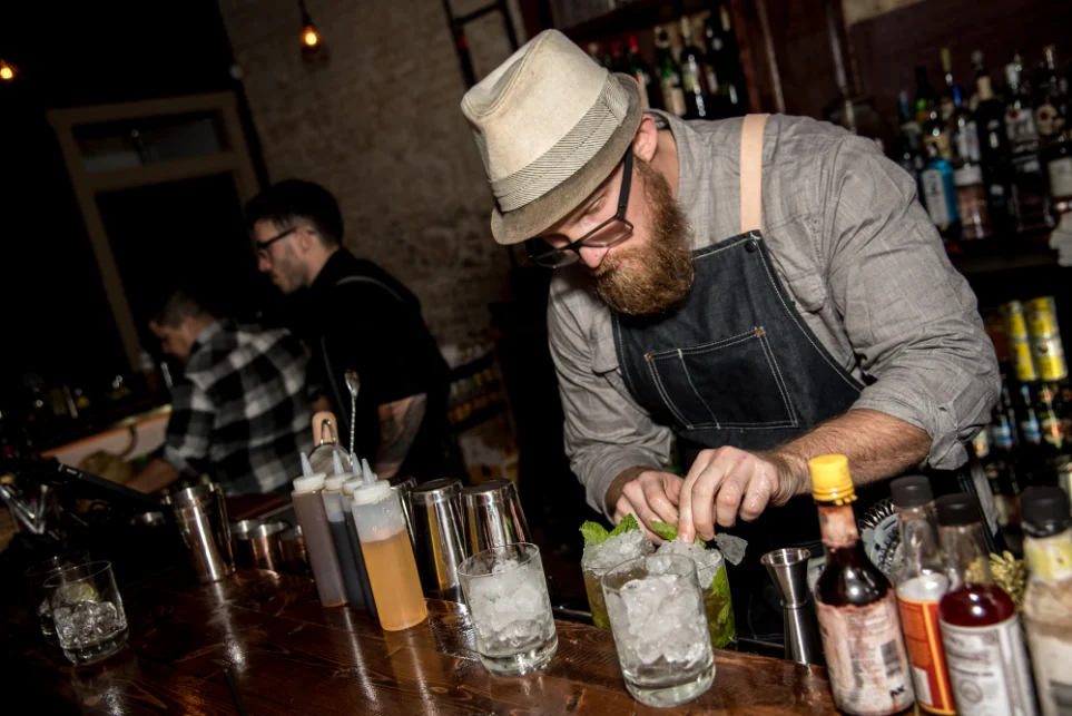 Bartender crafting a cocktail at a club in Dallas, focused and surrounded by various ingredients and bar tools